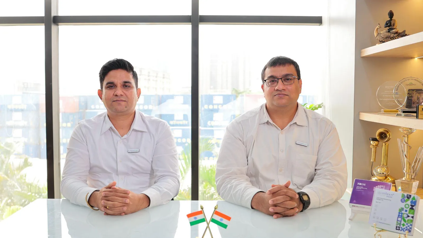 HaystackAnalytics co-founders seated at their office desk, with crossed Indian flags on the table and awards on the shelf behind them